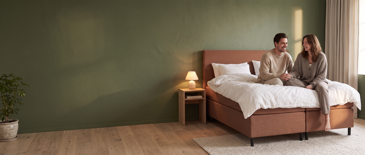 Jong stel zit lachend op een boxspring in Brown sugar-tint in een slaapkamer met een groene muur